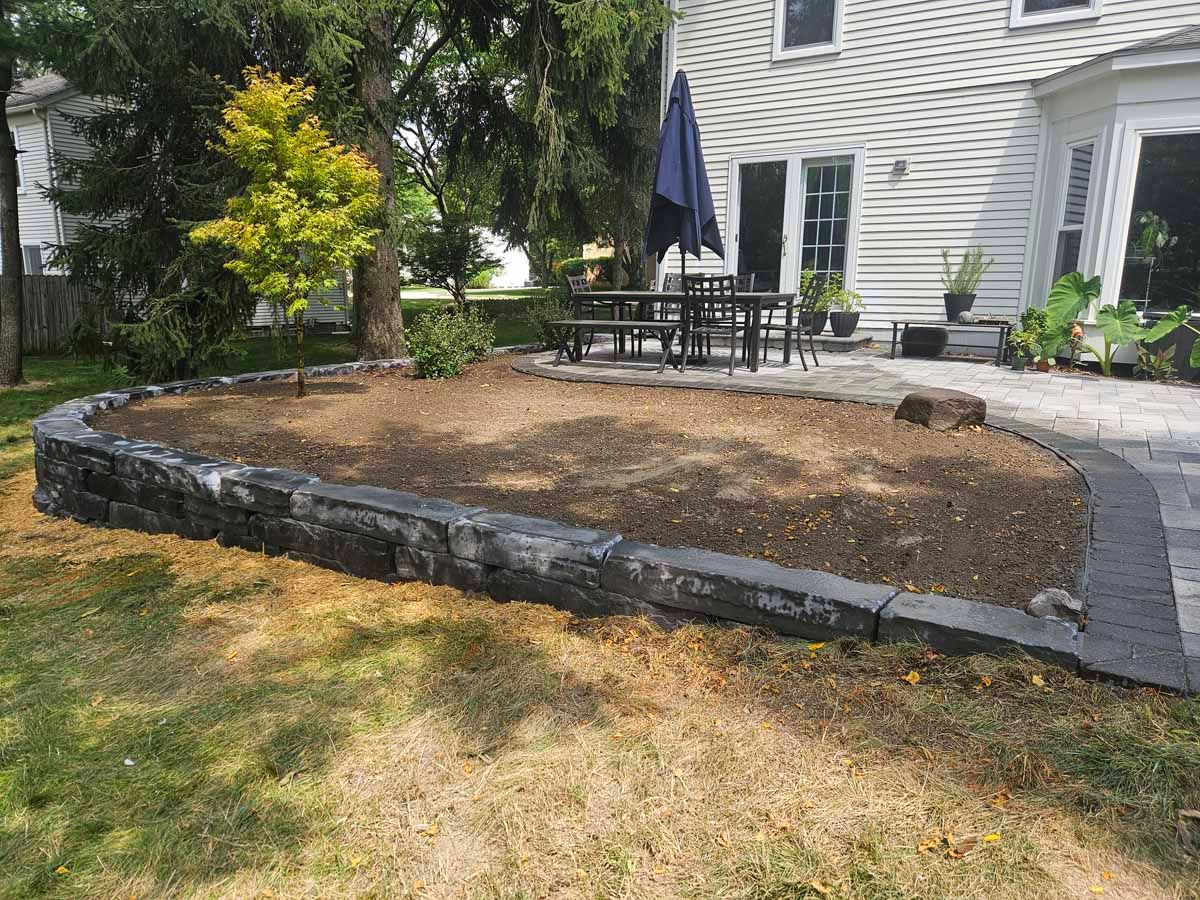 Raised stone garden bed in front of a white house with patio furniture and a small tree