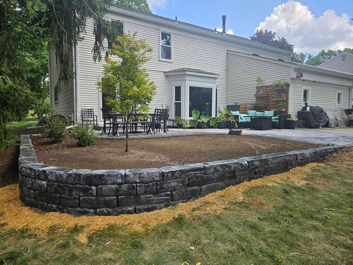 Raised stone patio beside a white house, with outdoor seating and a landscaped retaining wall