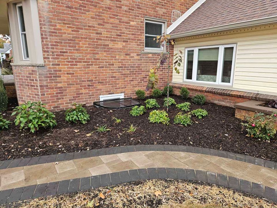 Curved garden bed with dark mulch and small shrubs beside a brick house and white-trimmed windows.