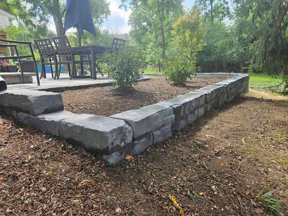 Stone retaining wall around a landscaped garden bed beside an outdoor patio and chairs.