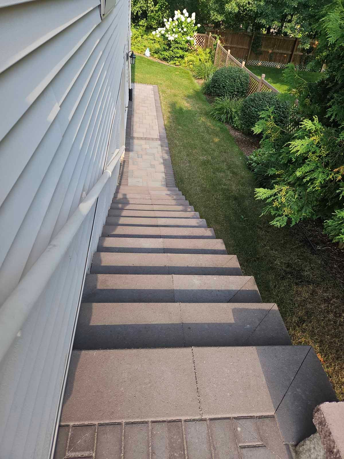 Narrow outdoor stairway beside white siding, descending past a grassy yard and shrubs
