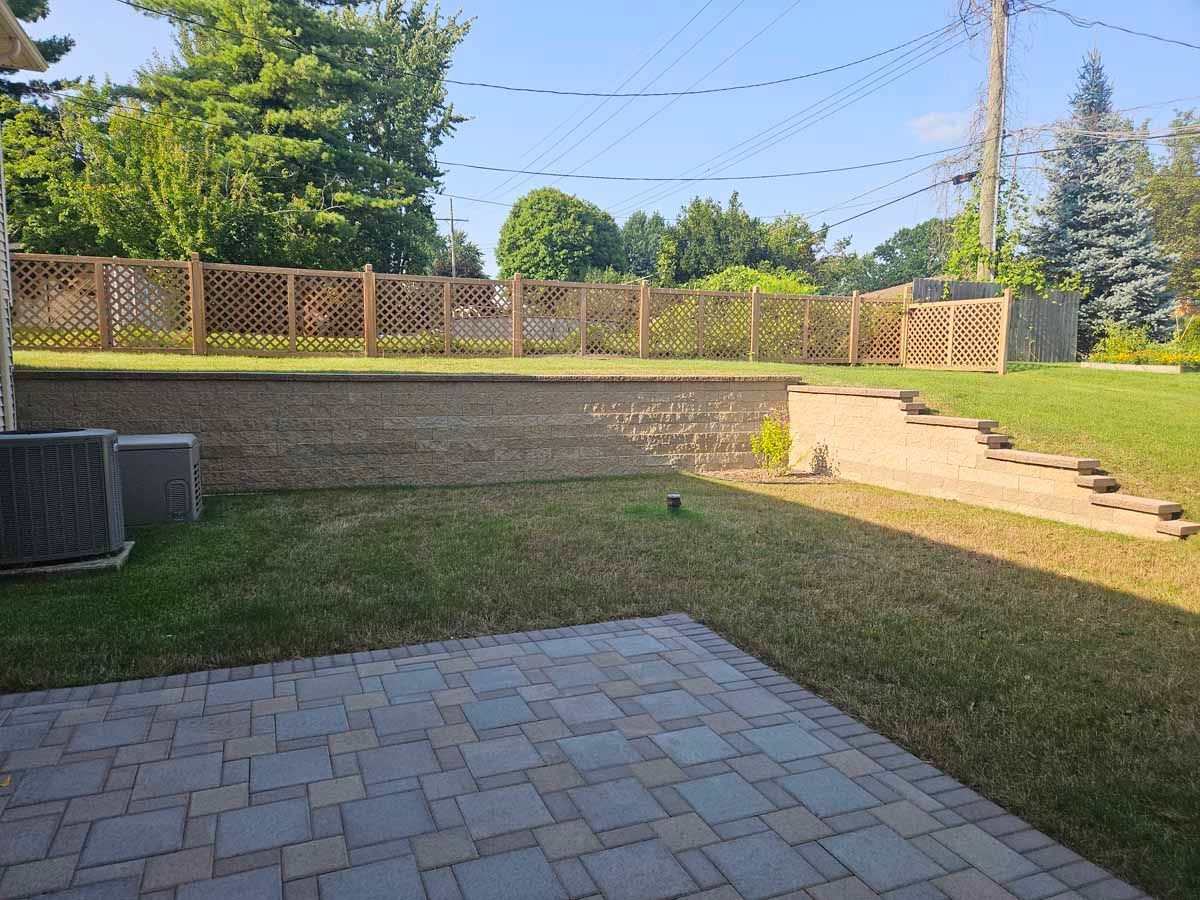 Backyard patio with stone pavers, grassy lawn, wooden fence, and raised garden bed in bright sunlight