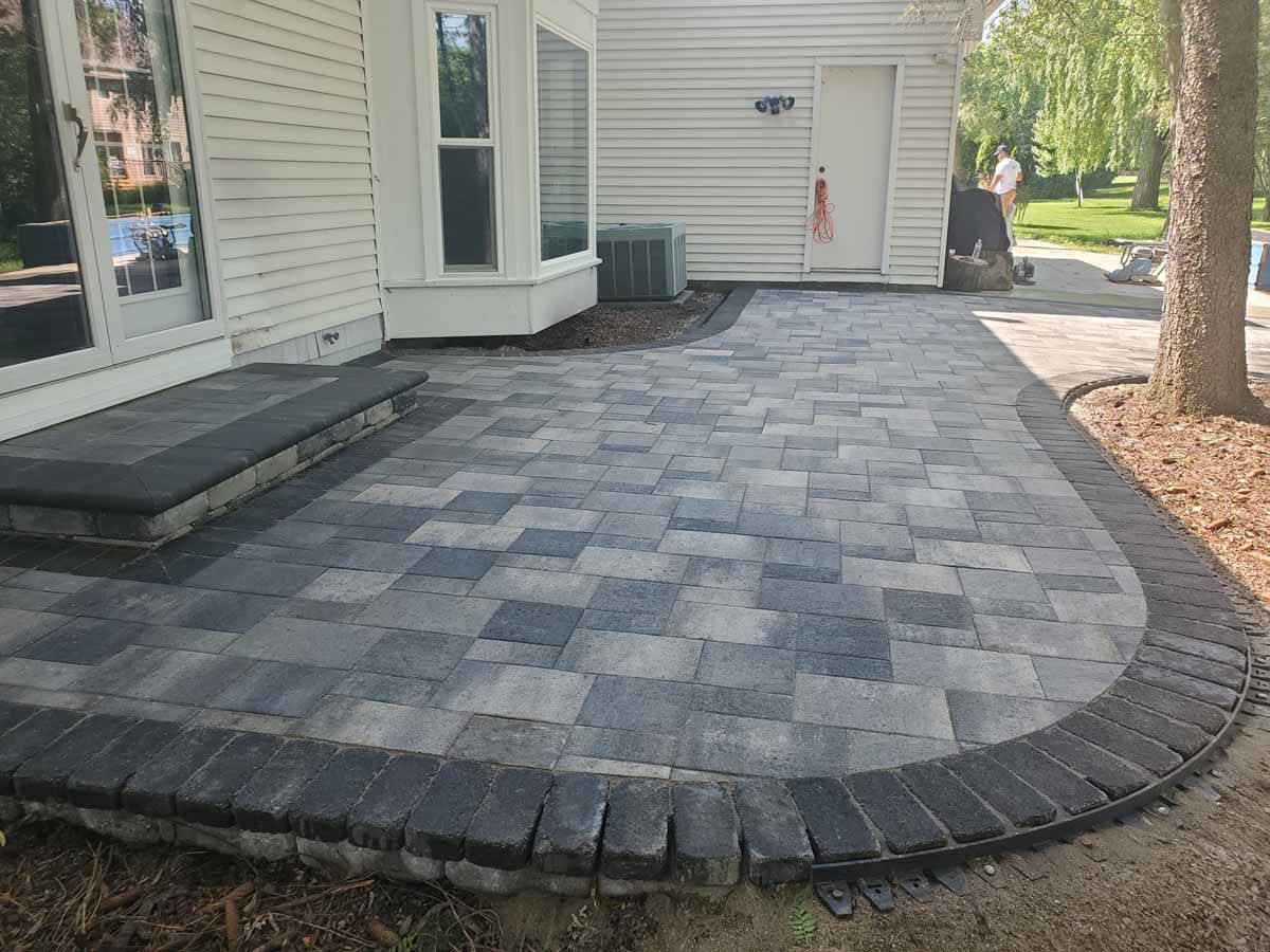 Curved gray paver patio beside a white house with a small raised step and bordered planting bed