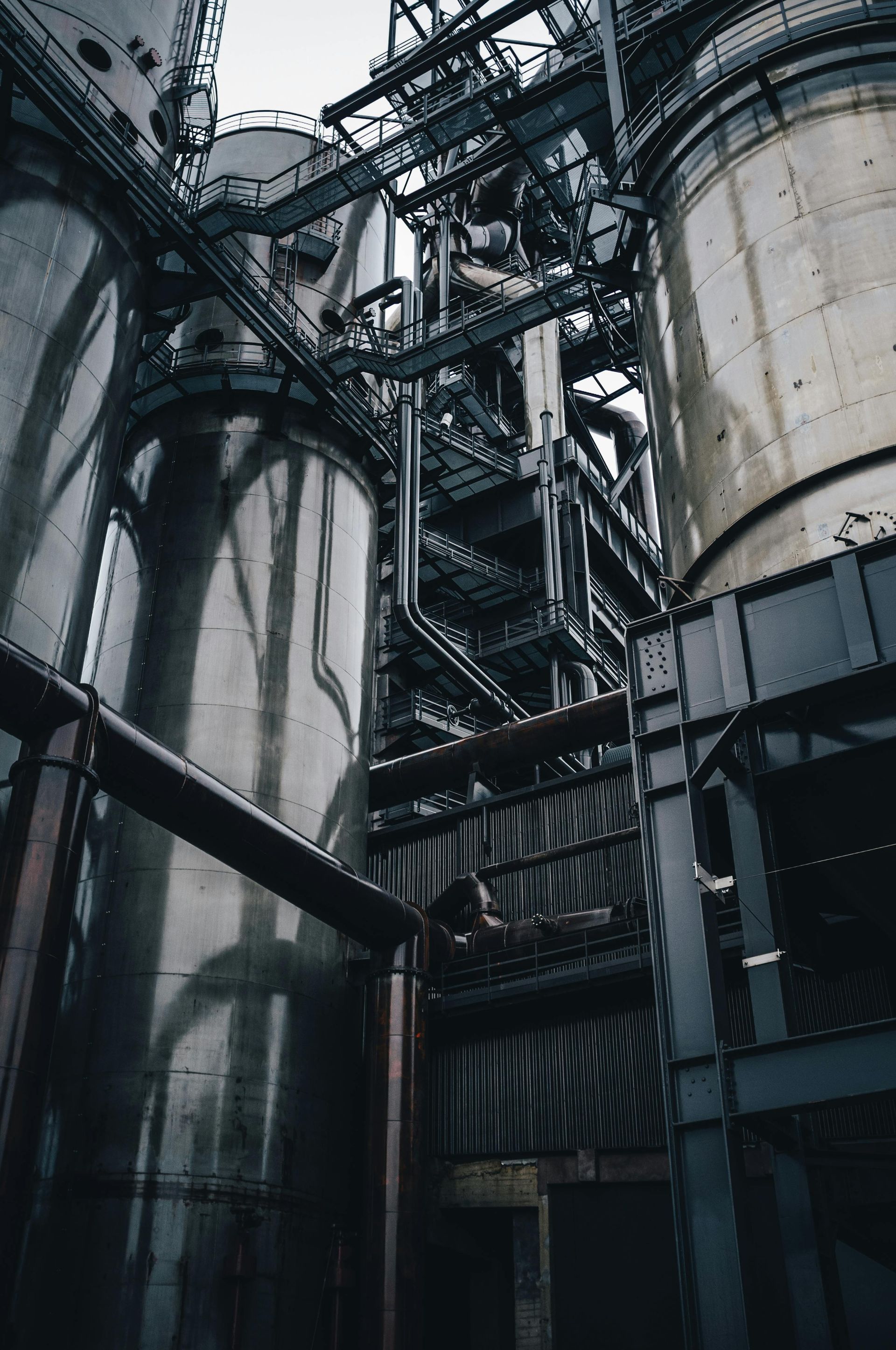 Industrial plant interior with tall metal silos, catwalks, and pipes in a dim, steel-blue setting