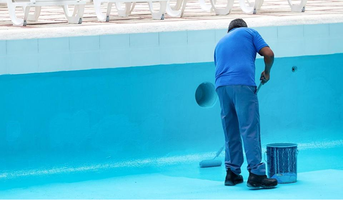 Worker in blue uniform cleaning the side of an empty swimming pool with a bucket nearby