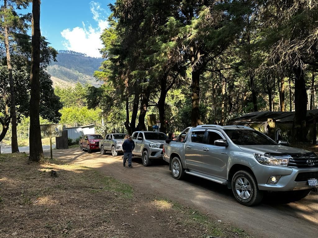 Todoterrenos aparcados en un camino de tierra en una zona montañosa boscosa, con gente cerca y tiendas de campaña al fondo.