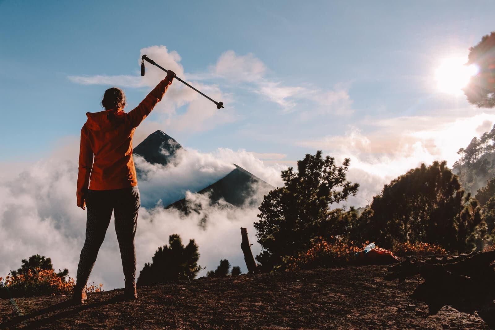 Un excursionista con chaqueta roja levanta un piolet en la cresta de una montaña al amanecer, con nubes y árboles debajo.
