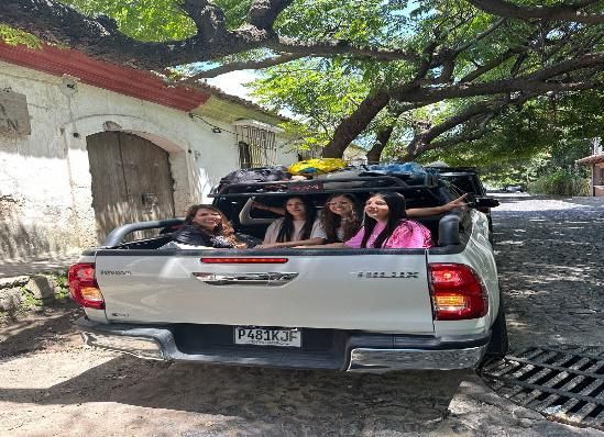 Gente viajando en la parte trasera de una camioneta plateada en una carretera bordeada de árboles.