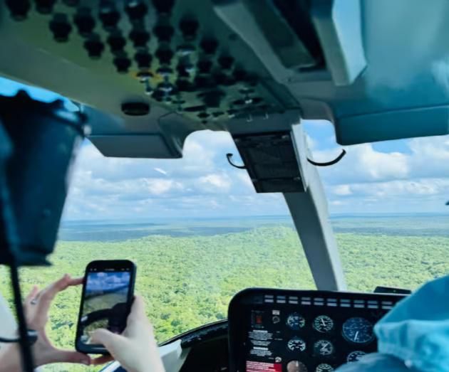 Dentro de la cabina de un helicóptero, un pasajero filma un denso bosque verde a través de la ventana delantera.