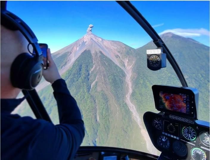 Vista desde la cabina de un helicóptero de una persona volando hacia un pico volcánico verde bajo un cielo azul.