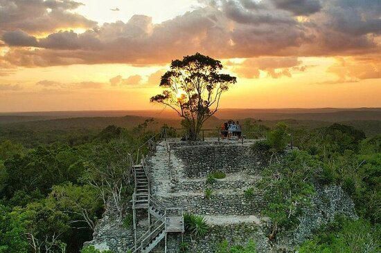 Puesta de sol sobre ruinas de piedra con un árbol solitario y gente en una terraza, cielo anaranjado brillante.