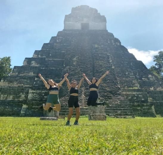 Tres personas posando con los brazos en alto frente a una pirámide de piedra escalonada bajo un cielo azul.