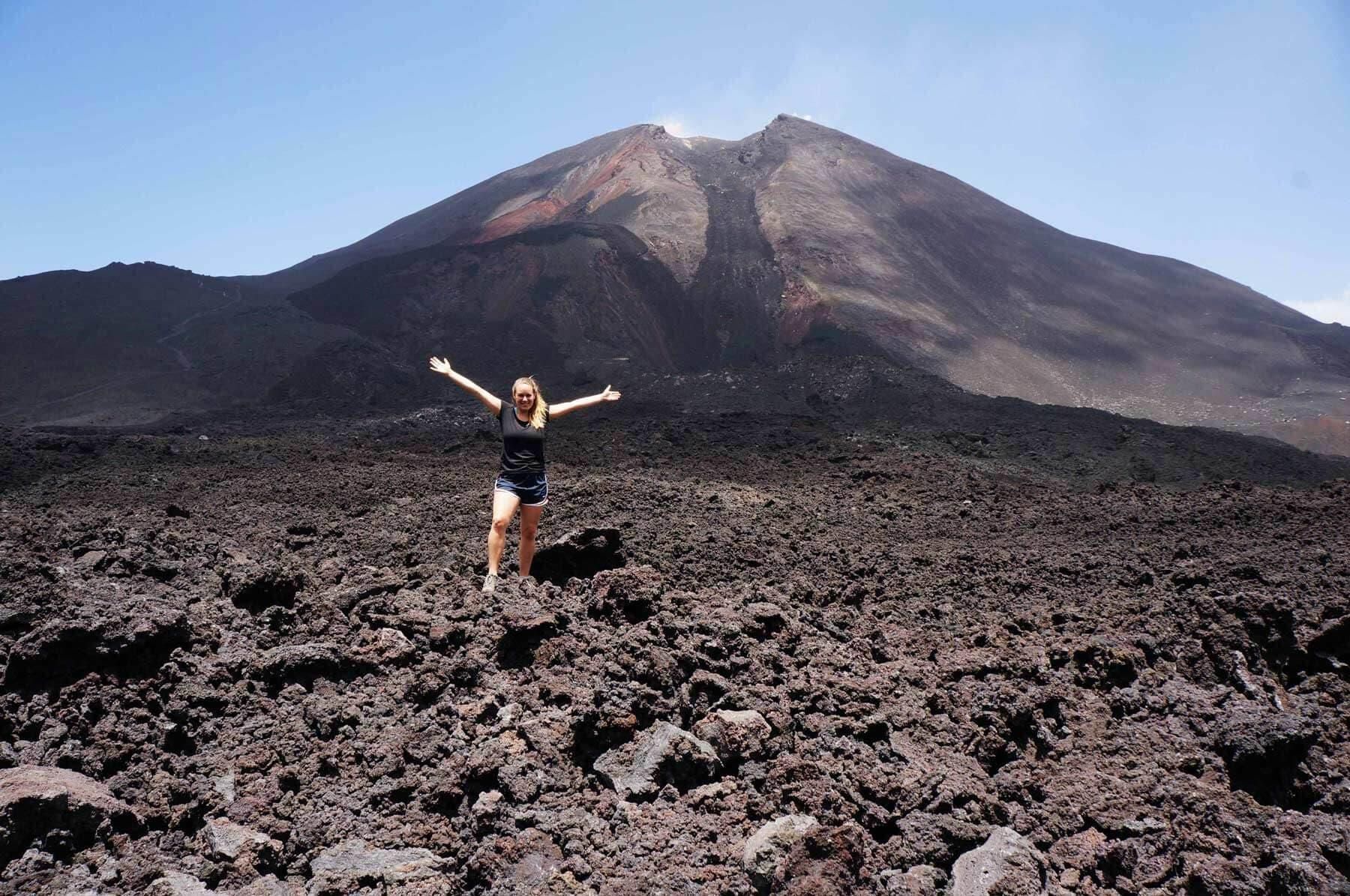 Persona de pie en un paisaje rocoso volcánico con los brazos en alto frente a una montaña.