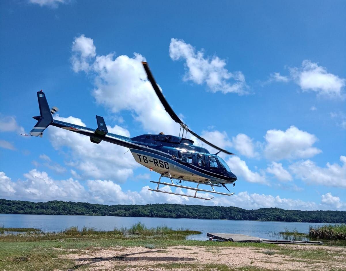 Helicóptero azul sobrevolando la orilla de un río bajo un cielo brillante con nubes.