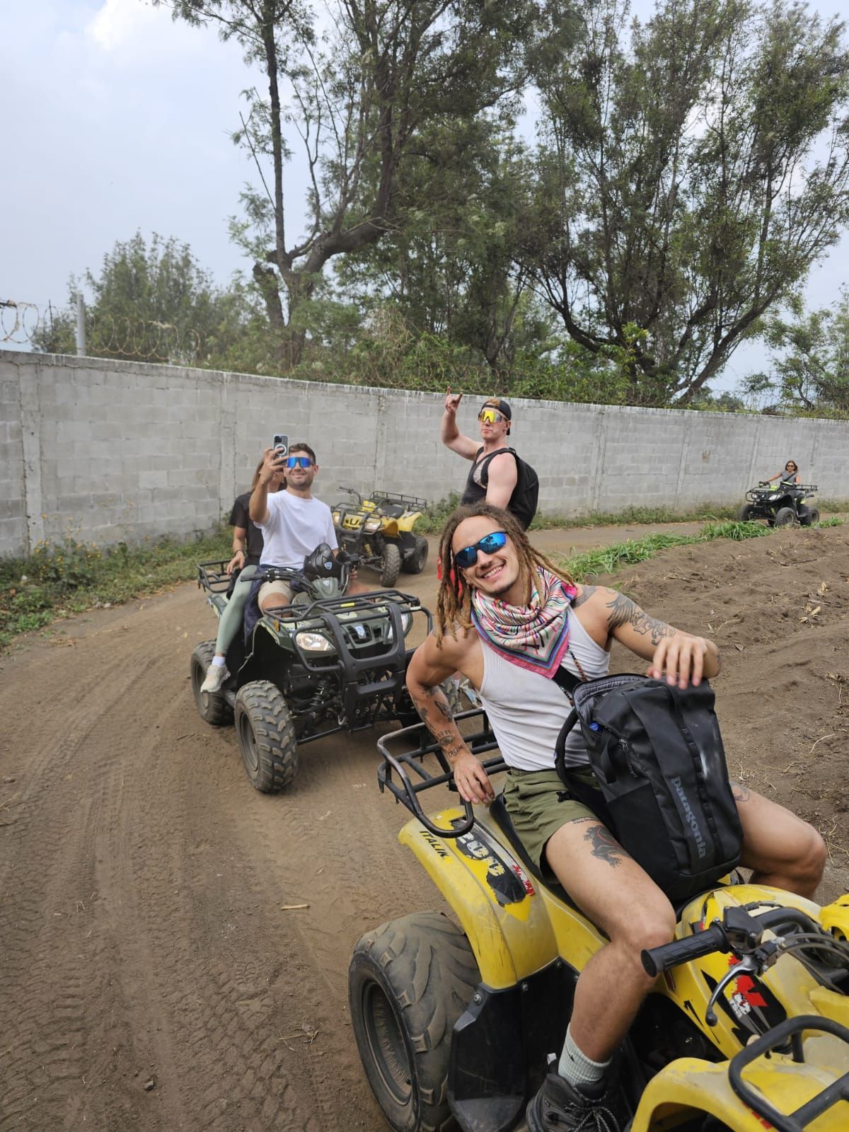 Gente montando quads en un camino de tierra junto a un muro de hormigón, sonriendo y saludando.