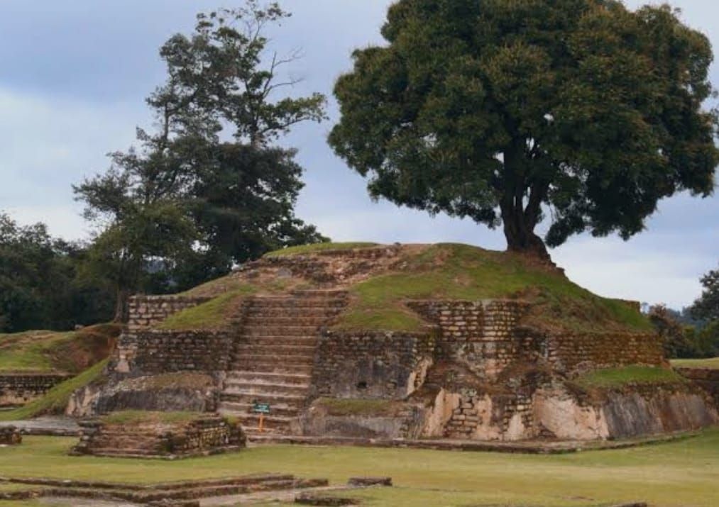 Ruinas de una antigua pirámide de piedra con escaleras, rodeadas de hierba y árboles bajo un cielo nublado.
