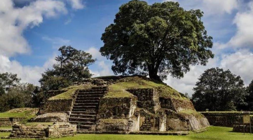 Ruinas de una antigua pirámide de piedra con escaleras, rodeadas de hierba y árboles bajo un cielo nublado.