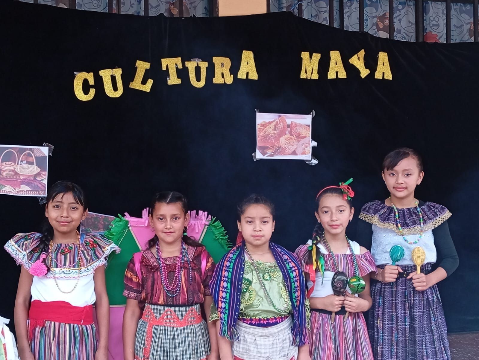 Cinco niños con coloridos trajes tradicionales están de pie en un escenario bajo una pancarta que dice “CULTURA MAYA”.