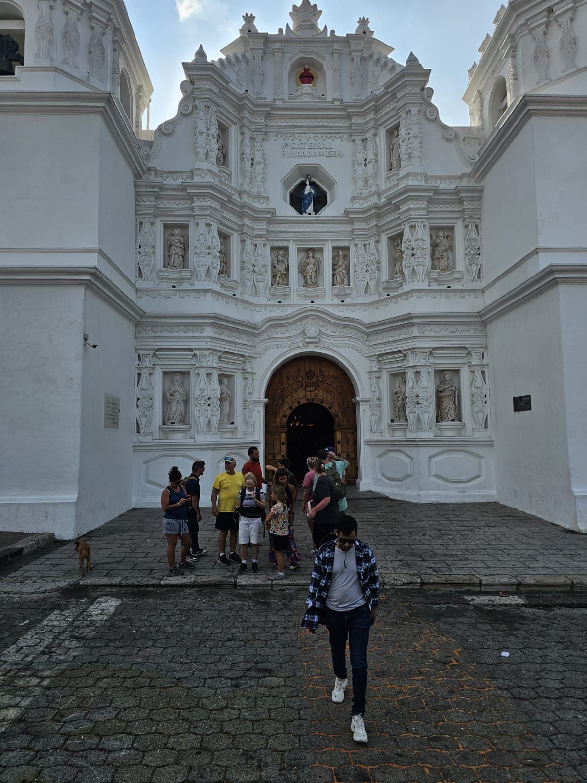 Turistas caminando hacia la ornamentada fachada blanca de una iglesia con una oscura entrada arqueada.