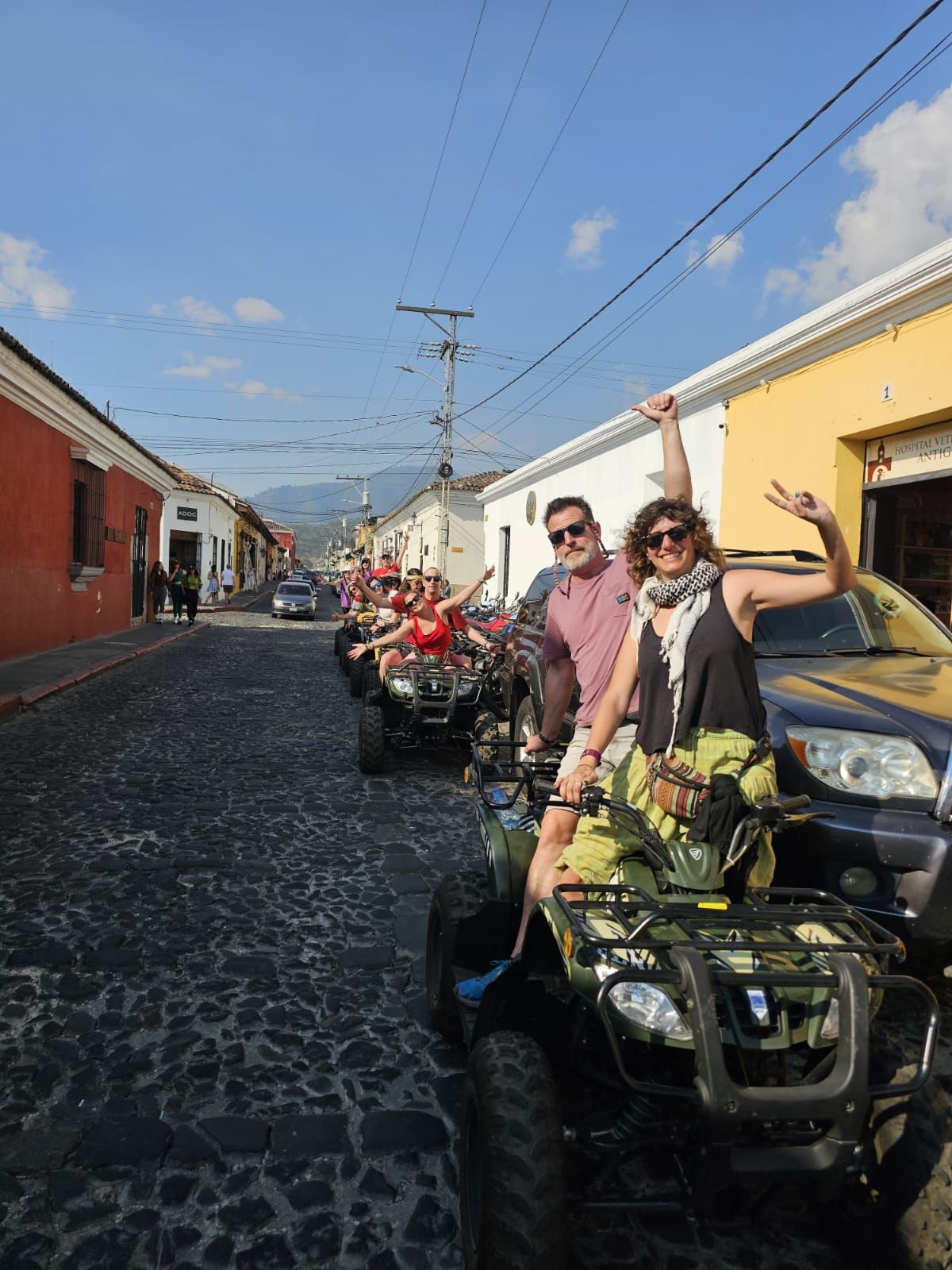 Dos personas pasean en un carrito de golf por una colorida calle empedrada con carritos estacionados