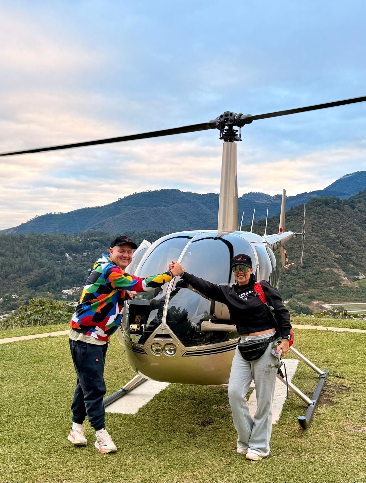 Un helicóptero azul vuela a baja altura sobre un lago cerca de una orilla cubierta de hierba bajo un cielo brillante.