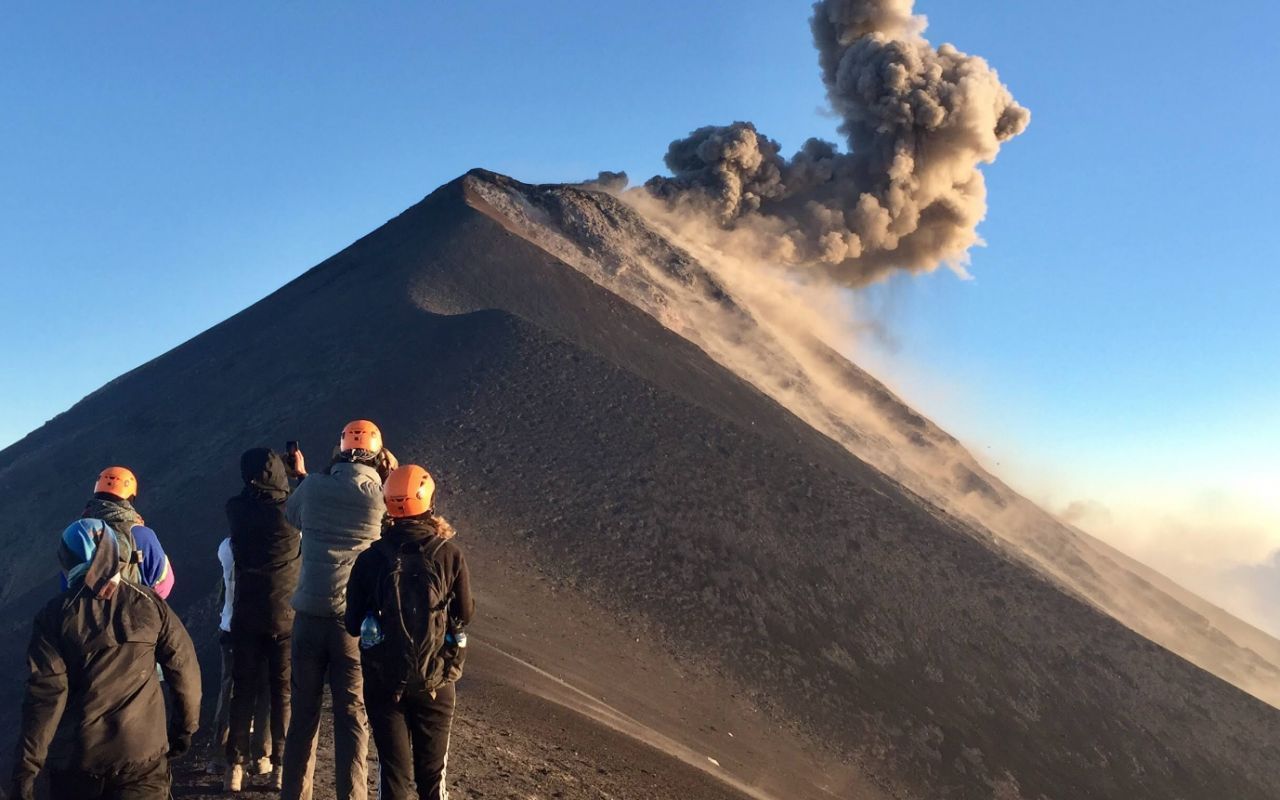 Un excursionista con chaqueta roja levanta un piolet en la cresta de una montaña al amanecer, con nubes y árboles debajo.
