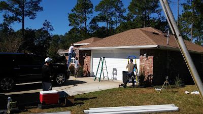 Workers install a white gutter system along the roofline of a brick house with a white garage door on a sunny day.