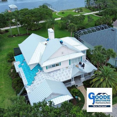 An aerial view of workers installing a light-colored metal roof on a house, featuring the Goode Brothers company logo.