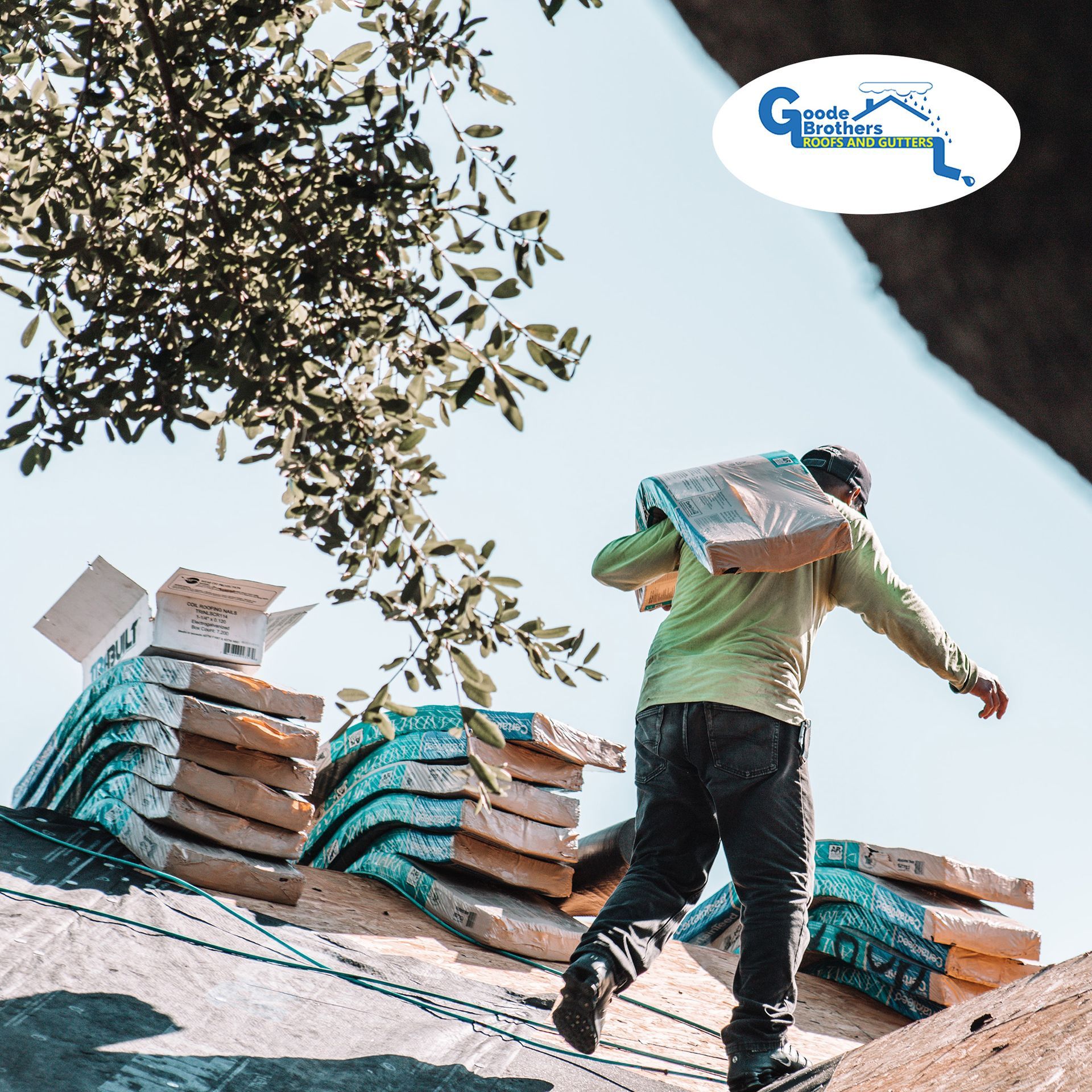 A worker in a long-sleeved shirt carries a stack of shingles across a residential roof under a clear blue sky.