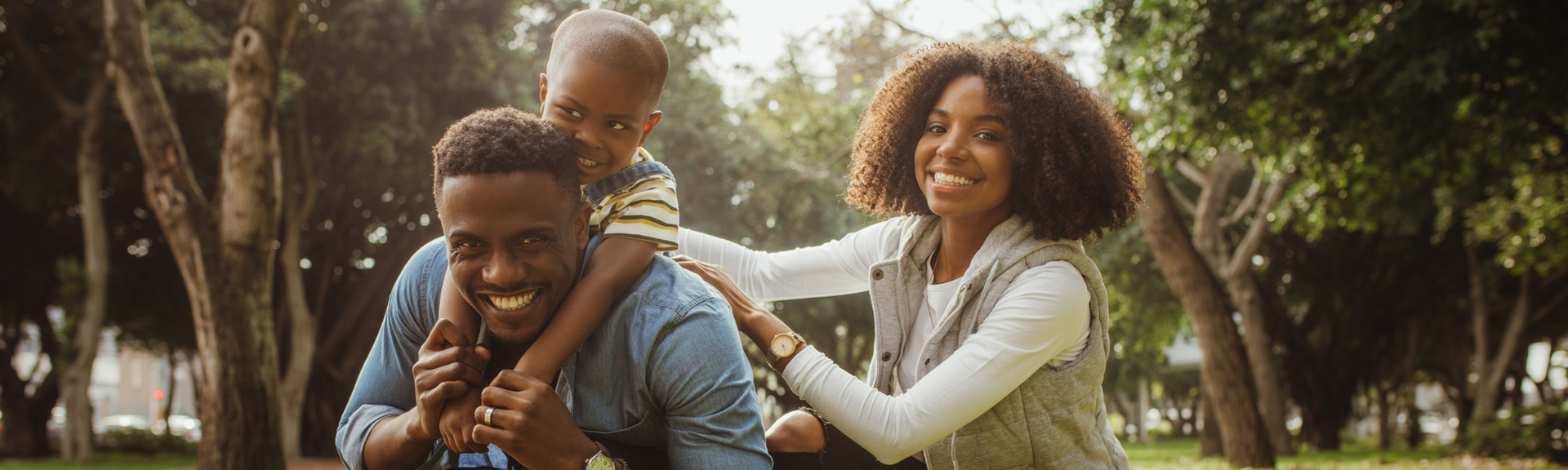 Family playing together in a sunlit park, smiling with a child on a parent’s back.