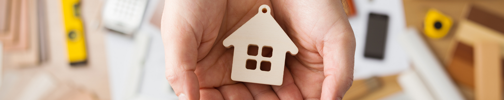 Hands holding a small wooden house in a bright home interior with blurred shelves in the background