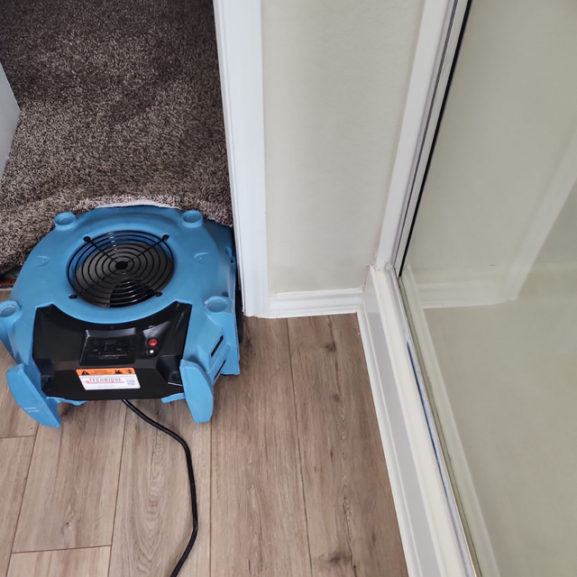 A blue commercial air mover fan positioned on a wooden floor, angled to dry carpeted stairs near a glass shower door.
