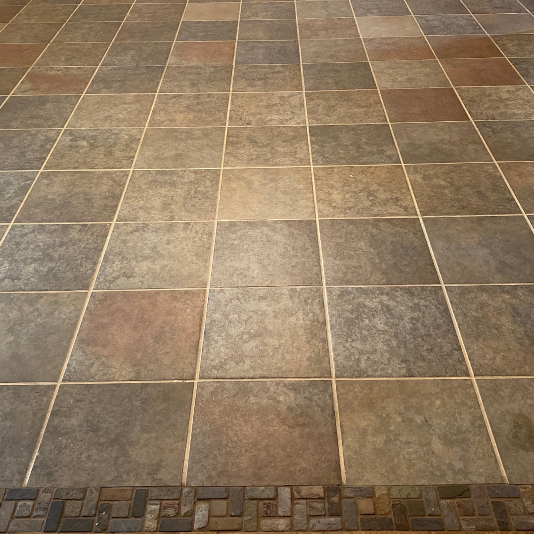 A floor made of square, multi-colored stone-look tiles with a border of small, rectangular dark bricks at the bottom.