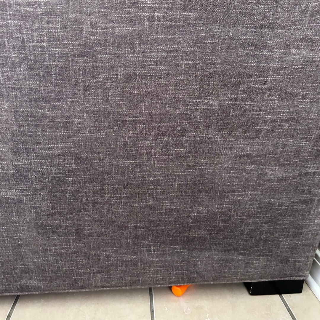 A gray fabric sofa sits on a tiled floor with a small, bright orange toy peeking out from underneath it.