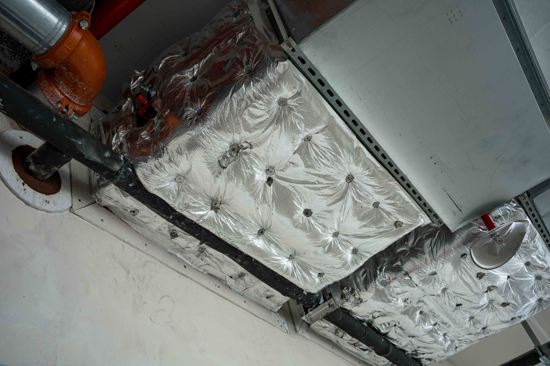 Insulated pipes and foil-wrapped ductwork on a ceiling, with a black metal support bar.