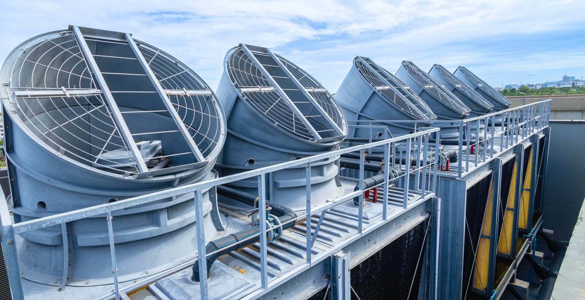 Row of large industrial cooling fans mounted on a rooftop, with metal railings and blue machinery below. Row of large industrial cooling fans mounted on a rooftop, with metal railings and blue machinery below.