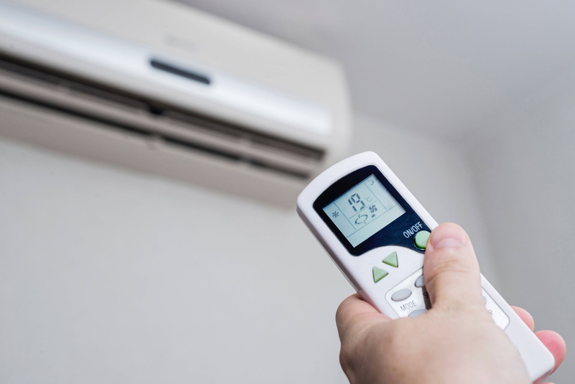 Hand holding remote aimed at wall-mounted air conditioner, adjusting temperature display.