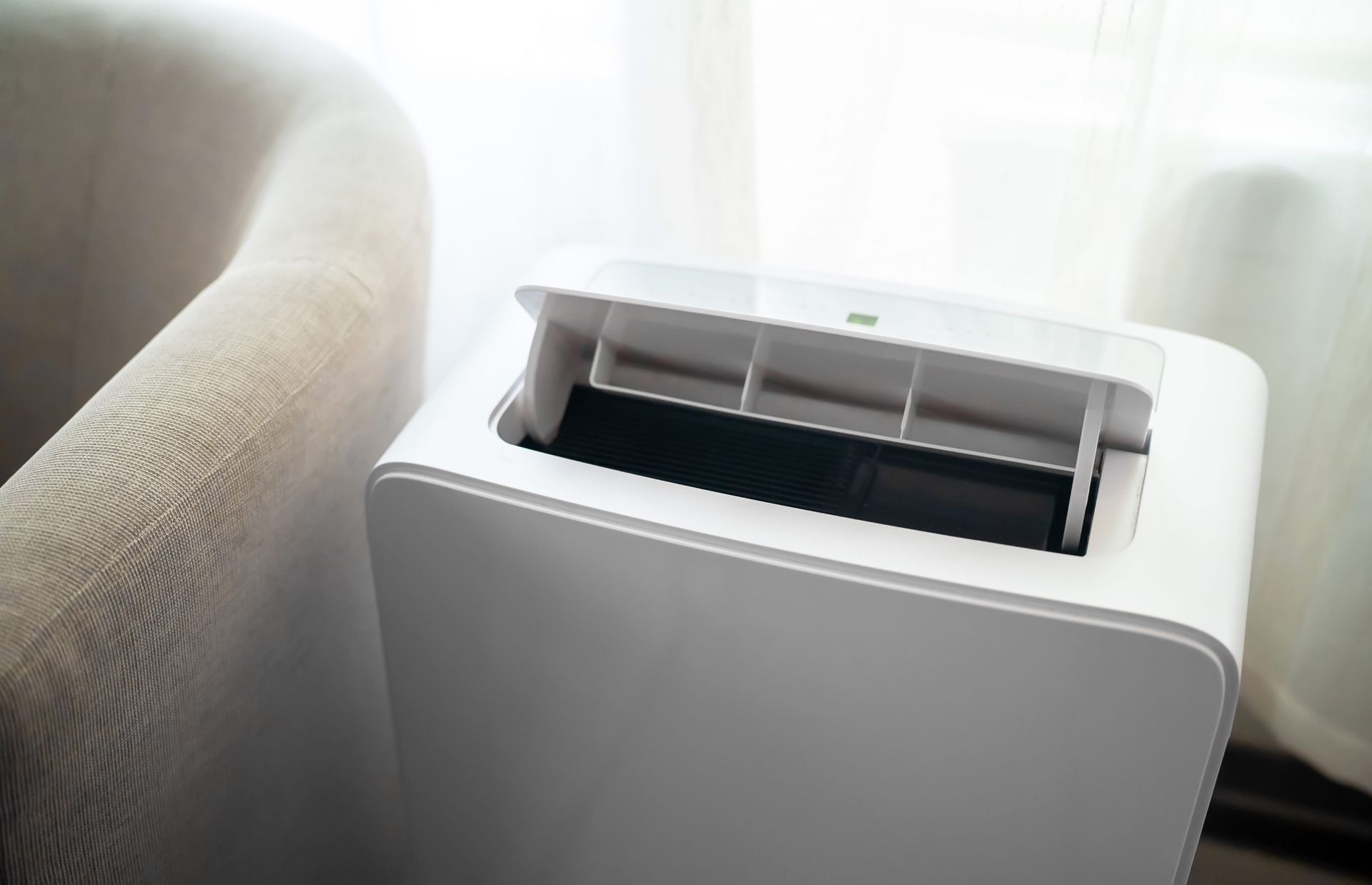 White portable air conditioner beside a beige couch near a sunlit window White portable air conditioner beside a beige couch near a sunlit window