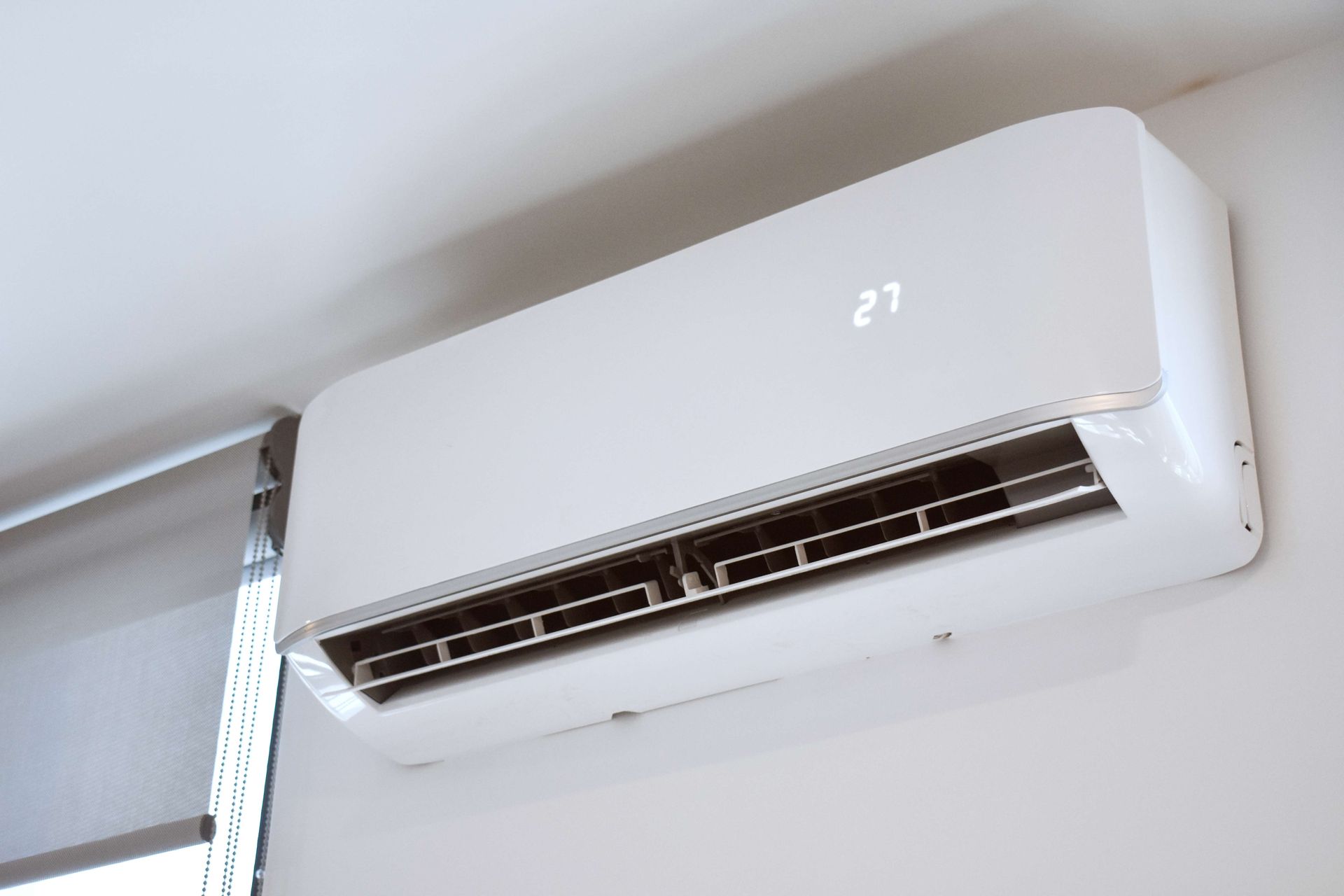 White wall-mounted air conditioner unit near the ceiling, with vents open and digital display showing 27.