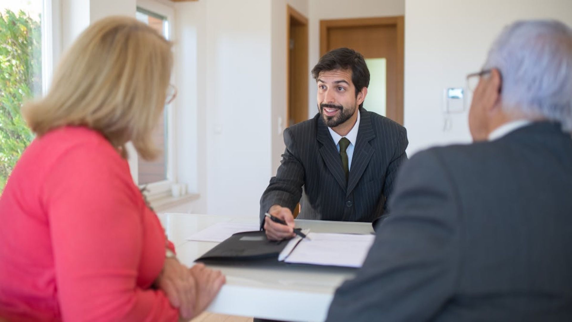 Hombre de traje hablando con una pareja mayor, revisando documentos en una mesa.