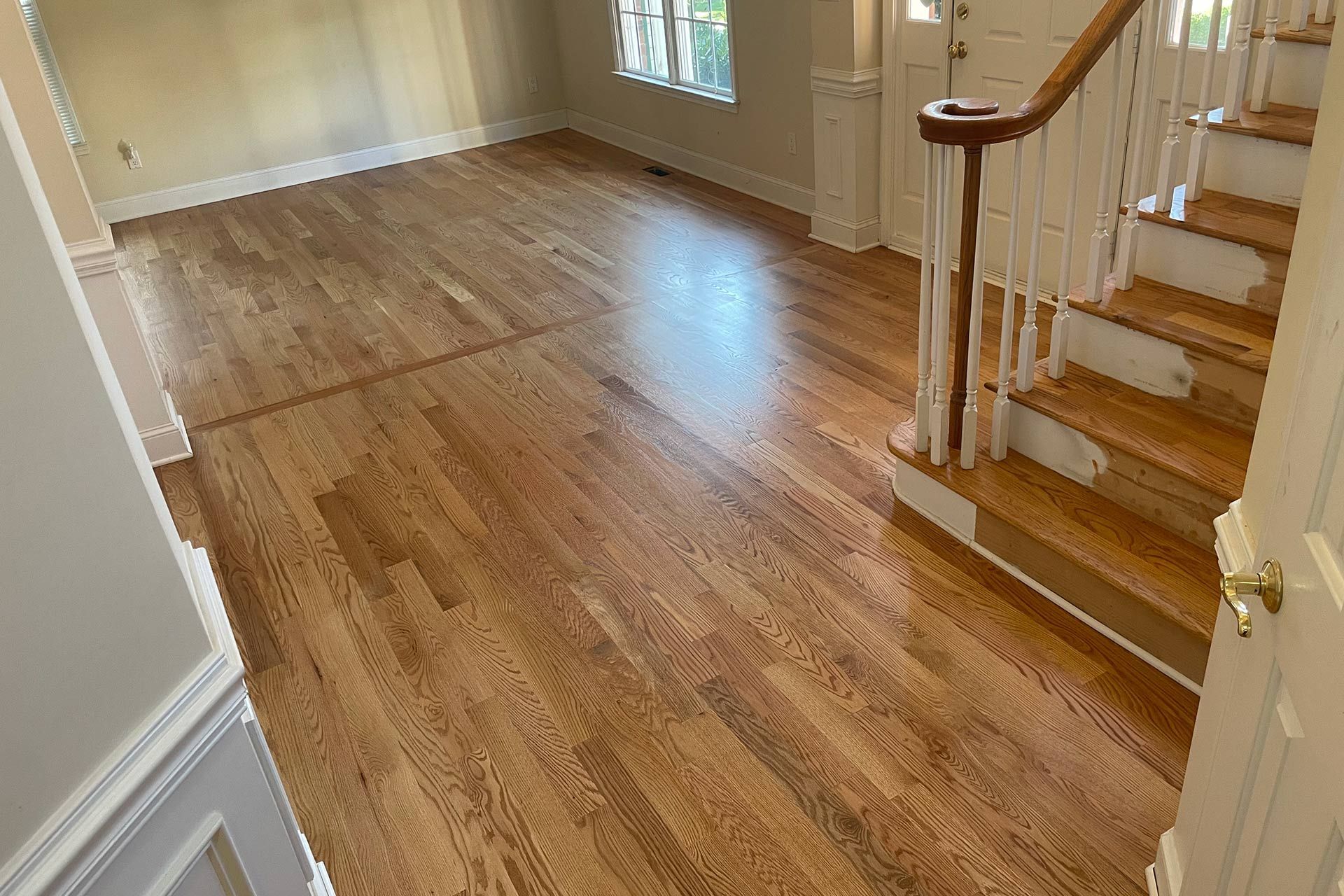Hardwood floor in home with stairs and doorway. Sunlight reflects on the polished wood.