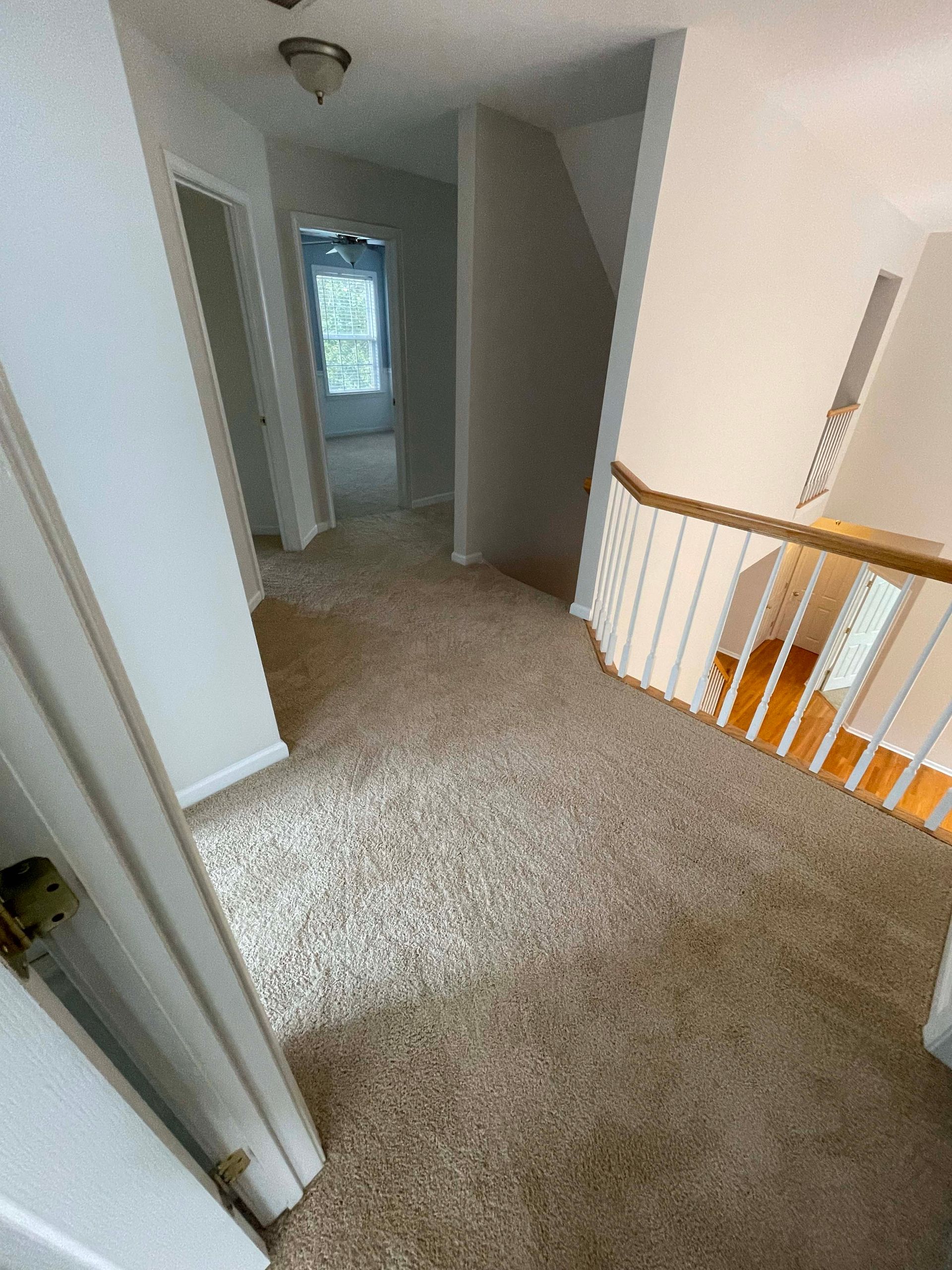 Hallway with tan carpet, white walls, and a staircase with a wooden railing.