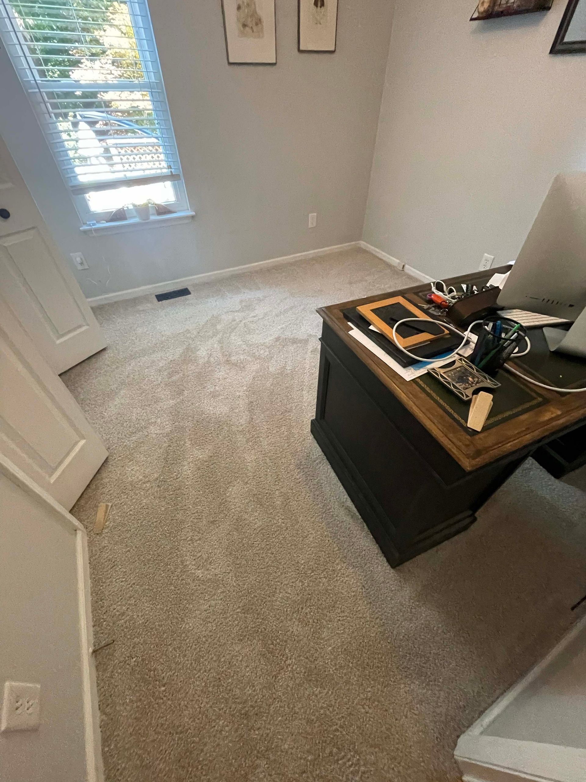 A room with beige carpet, a desk, a window, and two framed pictures on the wall.