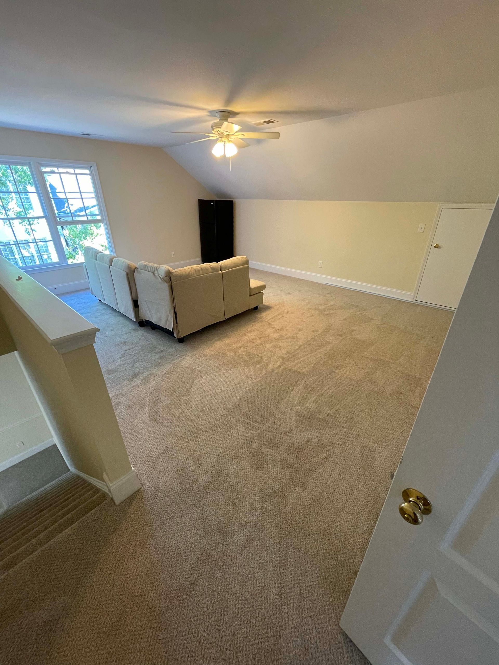Spacious carpeted room with a sofa, a window, and a door, likely an upstairs living area.