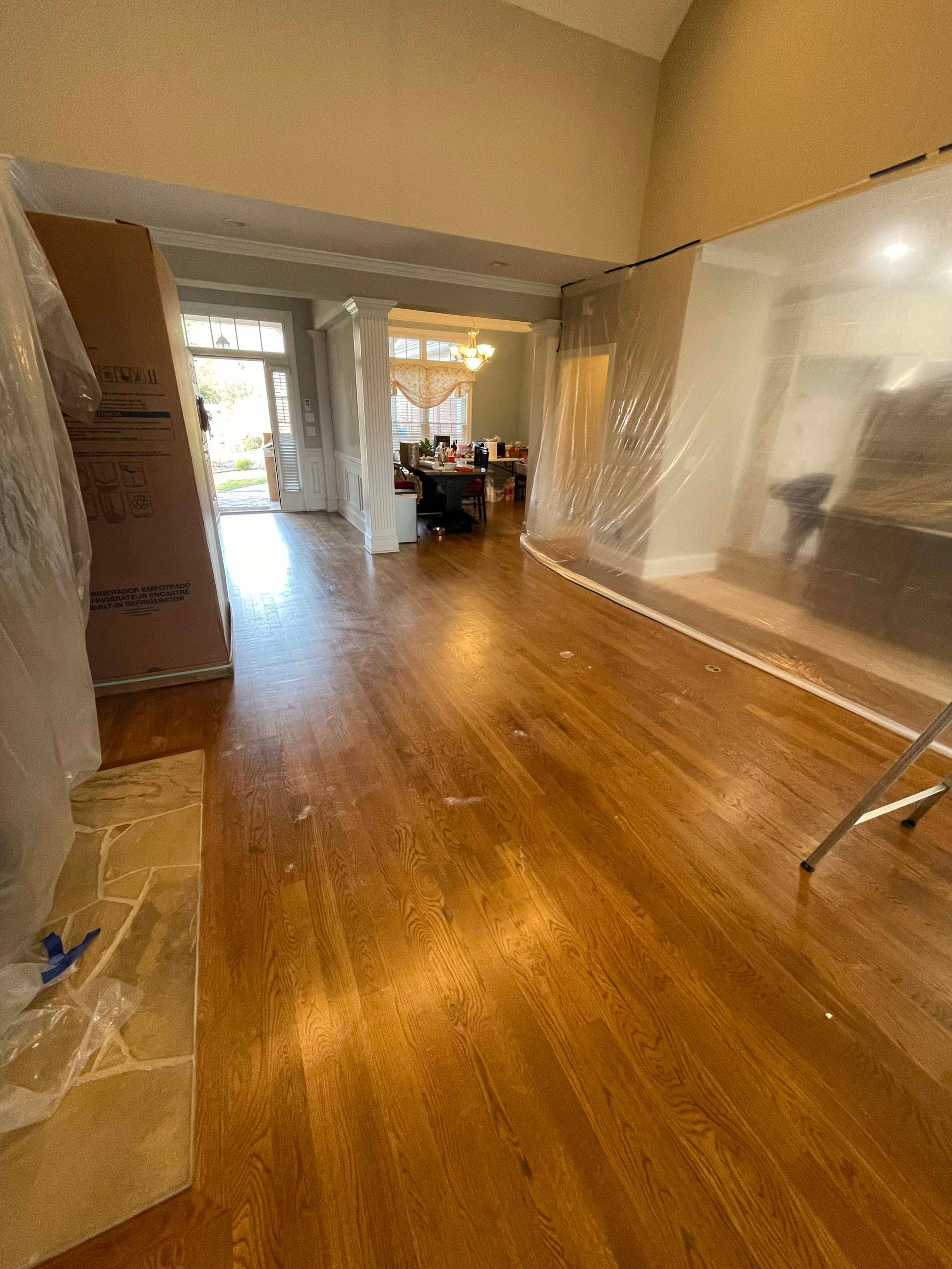 Interior view of a home under renovation with wood floors, protective plastic sheeting, and a distant dining area.