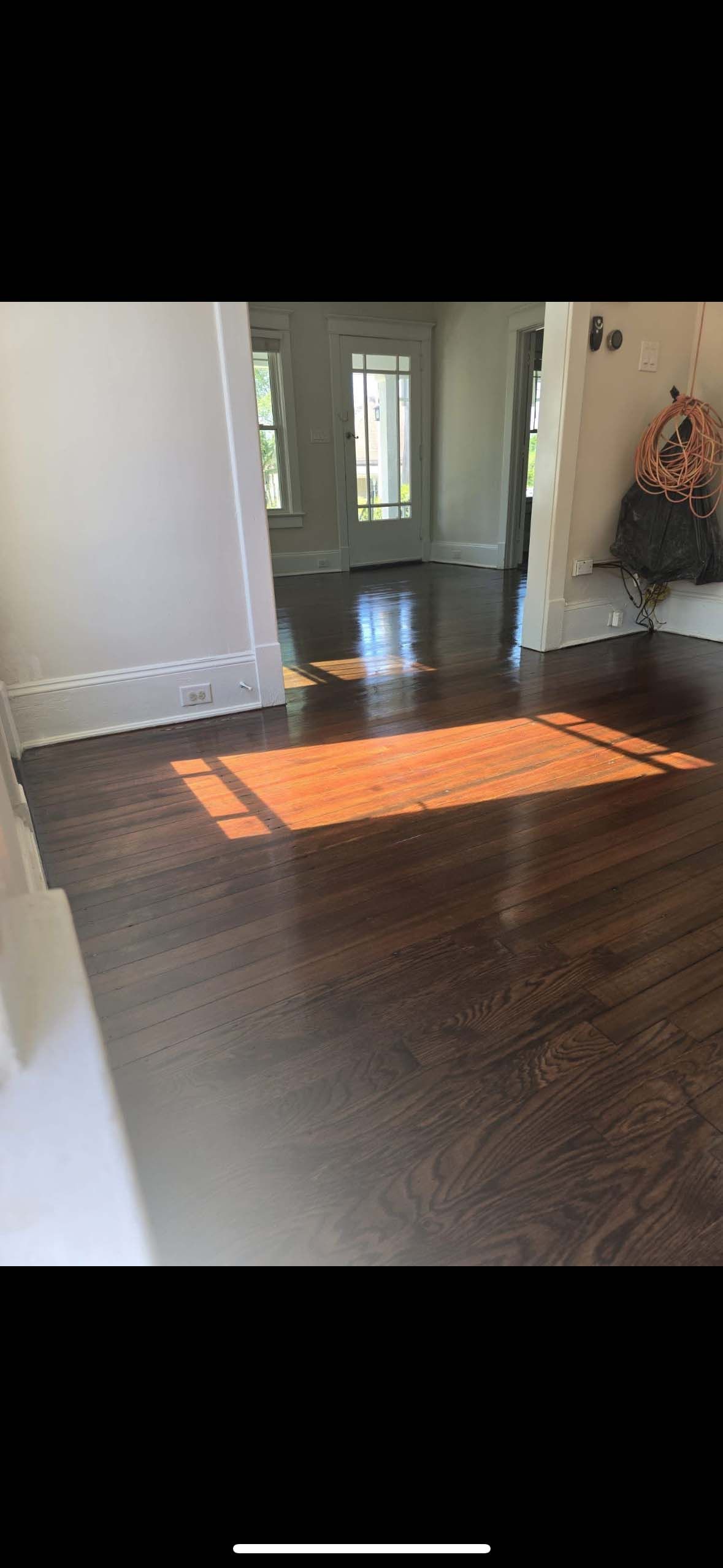 Sunlight illuminates a dark wood floor. Through a doorway, a hallway and a flower arrangement are visible.
