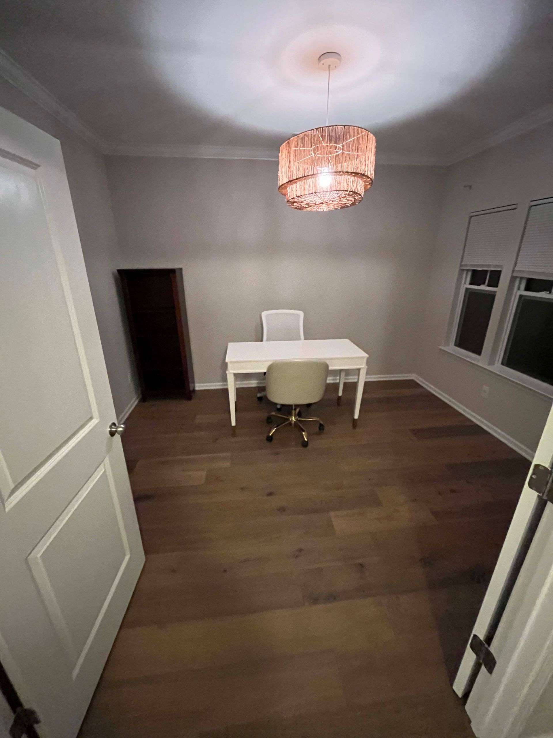 A home office with a white desk, chair, and a pendant light. Hardwood floors and white walls.