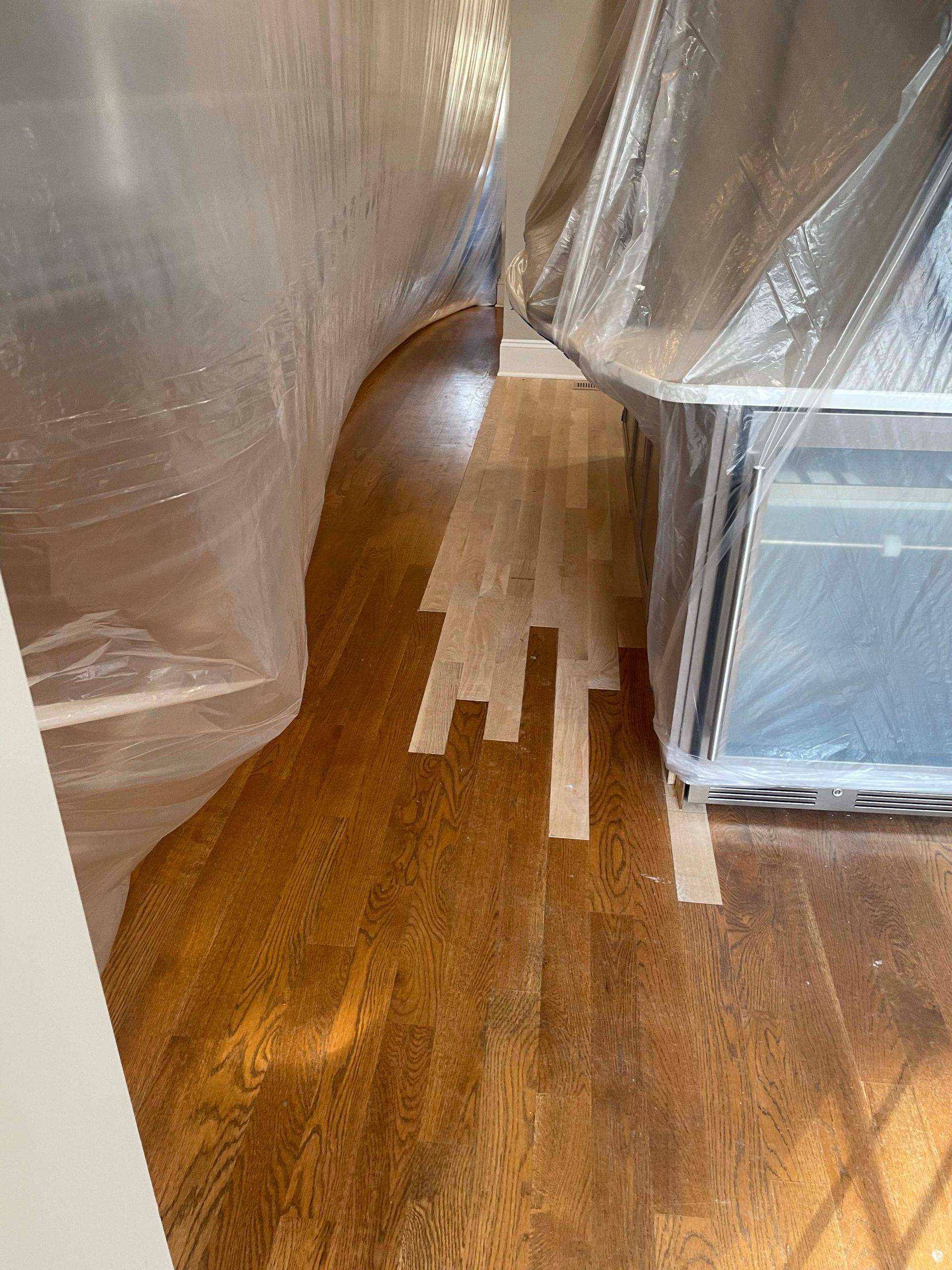 Wooden floor partially stained, protected by plastic sheeting. Tape marks the unfinished area.