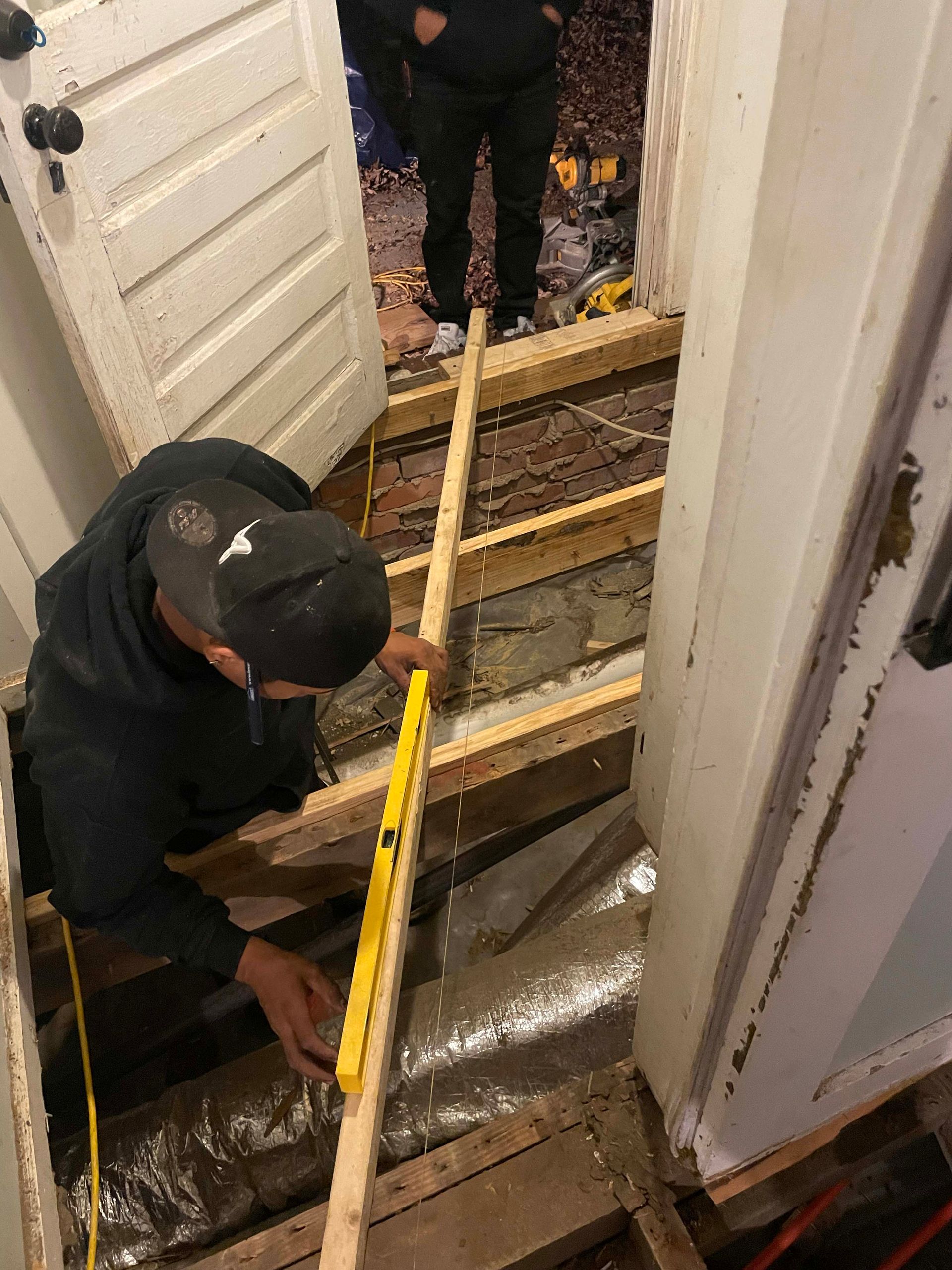 A person in a black hooded sweatshirt uses a yellow spirit level to check the alignment of a wooden floor joist.