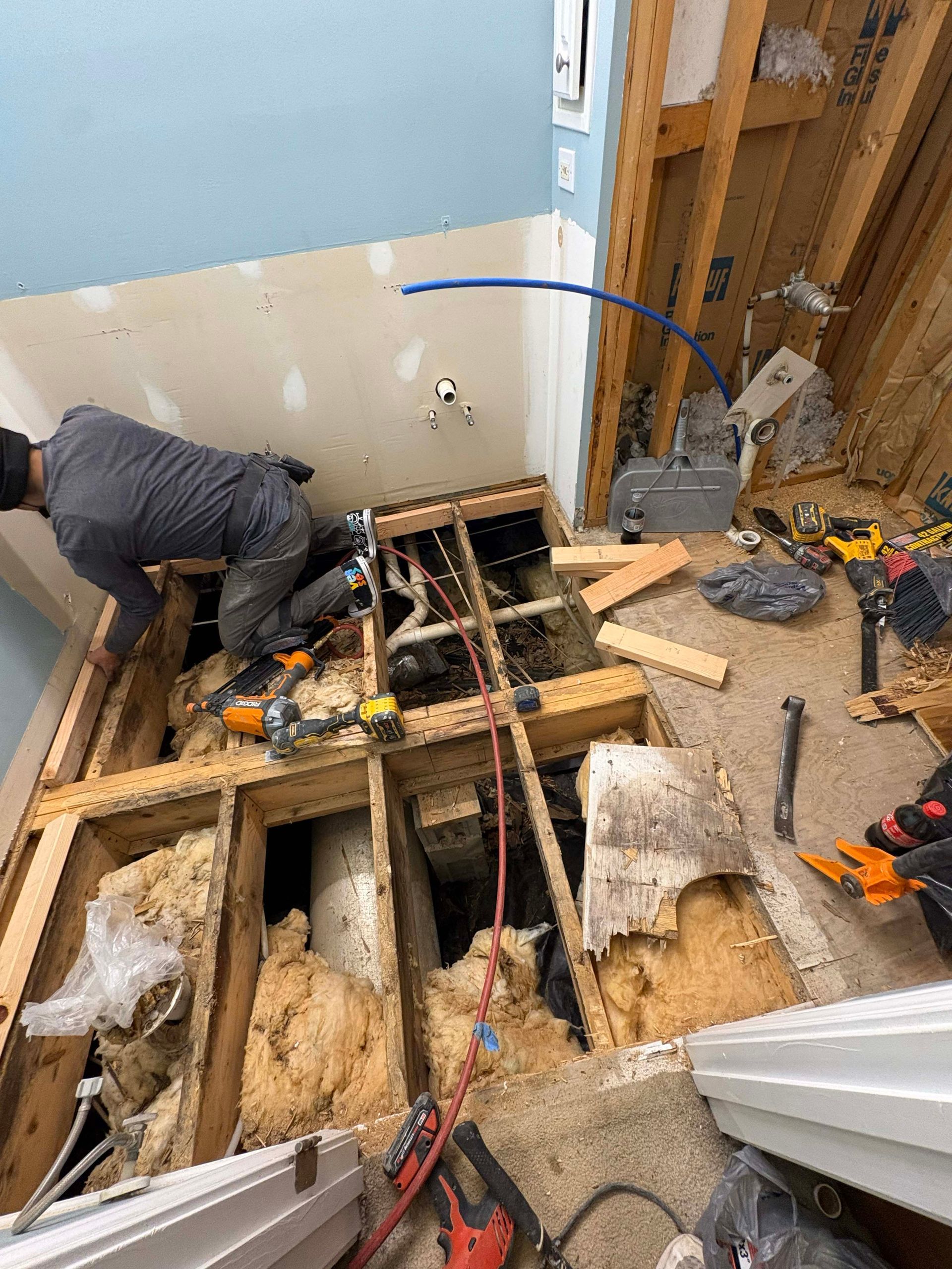 A worker kneels on a plywood floor, repairing plumbing pipes within an open subfloor frame inside a room under renovation.